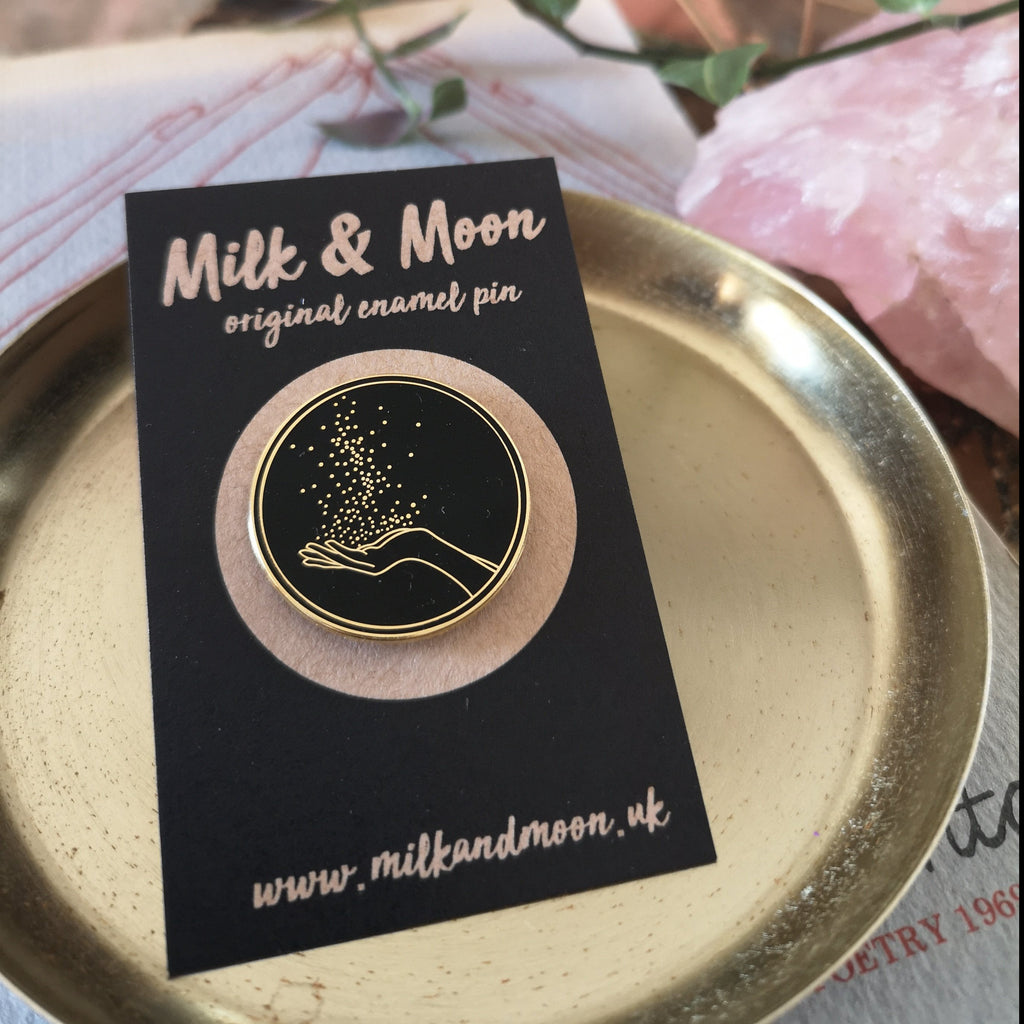 Milk and Moon Magic hard enamel pin , a black and gold circular enamel pin featuring an open hand in gold. There are sparkles rising from the open palm. 